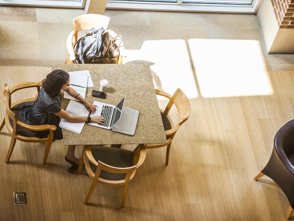 A female Online MBA student sits at a brown table working on her laptop while surrounded by papers and books